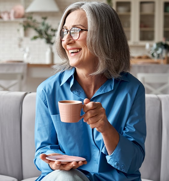 A middle-aged woman in a blue shirt smiling while holding a coffee mug, seated comfortably in a home-like setting.