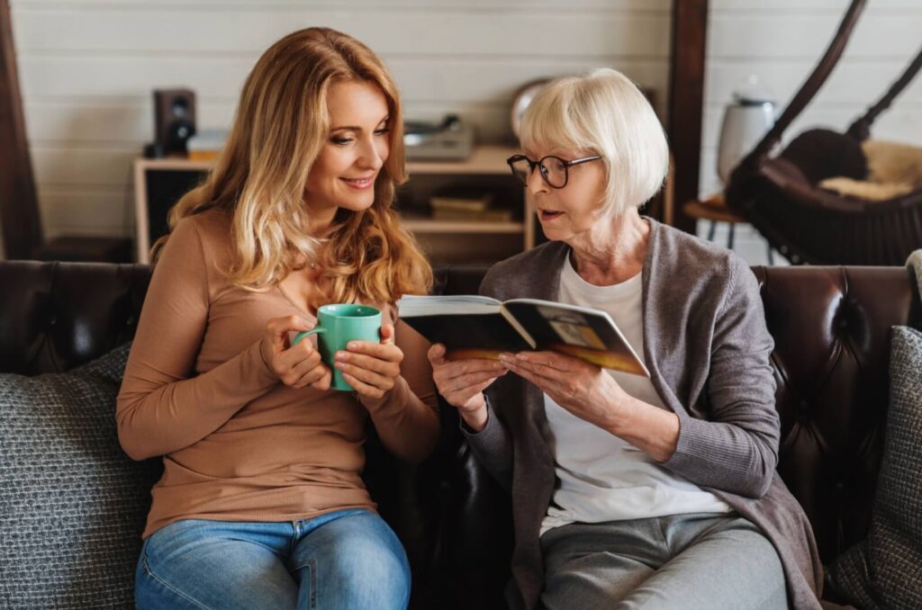 An older adult holding and looking at a photo album with their adult child sitting next to them.